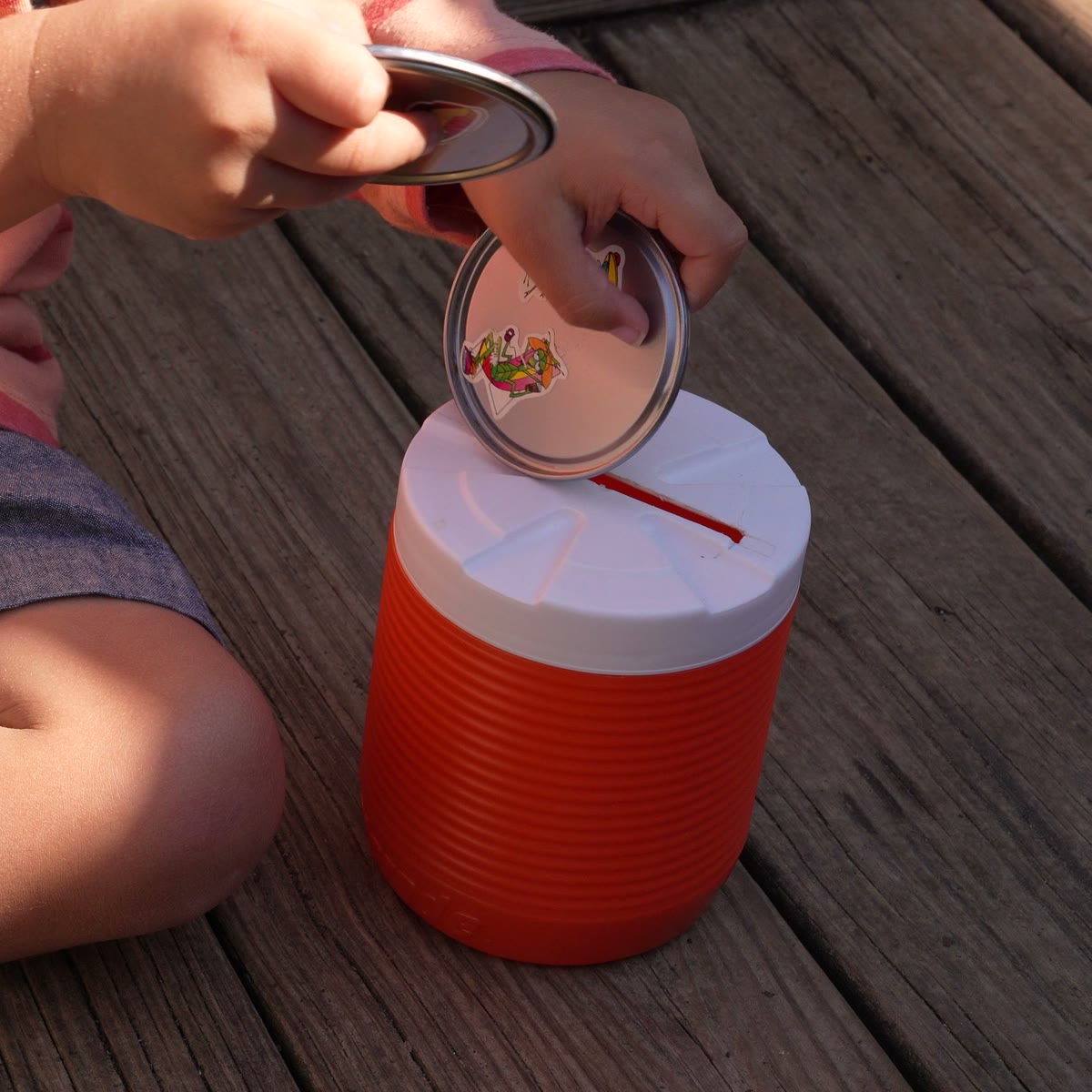 A child holding a jar lid against the slot in the top of the original orange canister, about to push it through.