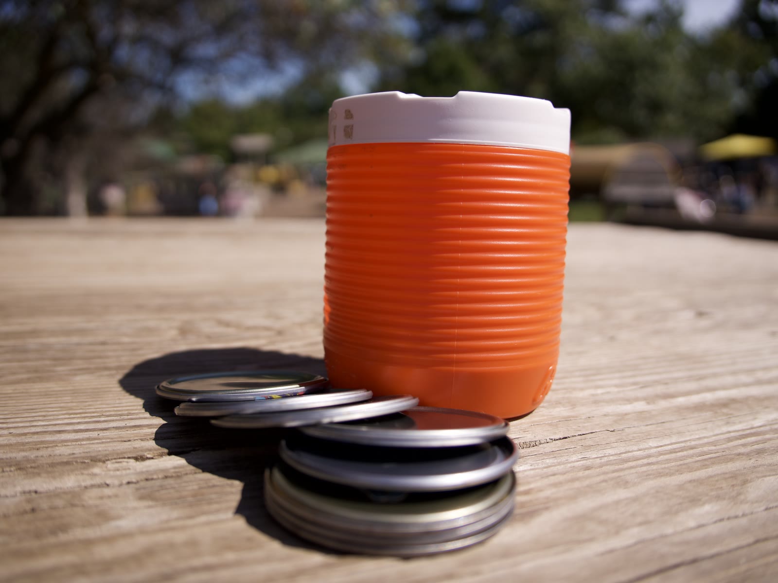 The original toy Martha made: an orange ribbed canister with a white screw top and a slot across it, a stack of jar lids leaning against it on a picnic table.